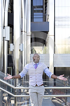 Elegant black enterprising man posing with his arms outstretched next to a luxurious office building. Business concept