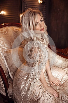 Elegant blond woman in beige dress posing on luxury sofa in royal interior. Fashion beautiful sensual bride with makeup, curly
