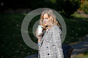 Elegant blonde with long curly hair in light tweed jacket drinking hot coffee from recyclable cup while walking on sunny