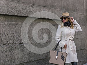 Elegant blonde woman with black glasses coat and hat walks down a street