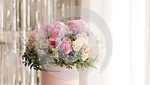 An elegant bouquet of fresh roses, peonies, and daisies in a pink box, beautifully arranged and placed by a light-filled