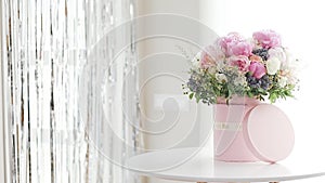 An elegant bouquet of fresh roses, peonies, and daisies in a pink box, beautifully arranged and placed by a light-filled