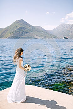 An elegant bride with a bouquet in her hands stands on a pier in the Bay of Kotor near the blue transparent water