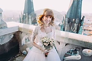 Elegant bride with bridal bouquet poses on the tower balcony of old gothic cathedral