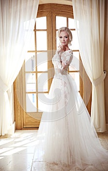 Elegant bride in fashion wedding dress. Blond woman model in white bridal gown with skirt posing in front of curtains window, ind