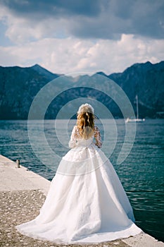 An elegant bride in wreath stands on a pier in the Bay of Kotor near the blue water, back view