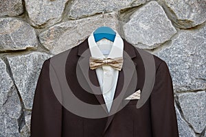 Elegant brown male tuxedo with white shirt and bow tie hanging on stone wall outdoors, copy space. Modern men accessories.
