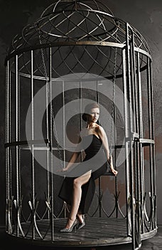 Elegant brunette woman in black dress sitting on a swing in big birdcage, like a symbol of isolation.