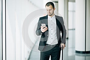 Elegant businessman checking e-mail on mobile phone while walking with suitcase inside airport terminal. Experienced male employer