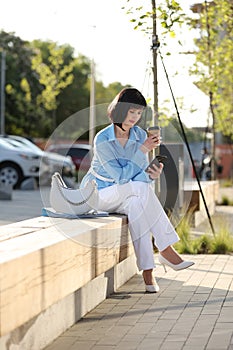 Elegant businesswoman holds paper cup with coffee or tea to go, messaging on smartphone and relaxing free time outside