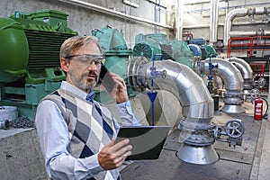 Elegant CEO Looking At Digital Tablet Screen And Talking On Smartphone Beside Large Generators And Pipes