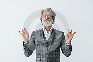 In elegant clothes. Standing against white wall. Senior stylish modern man with grey hair and beard indoors