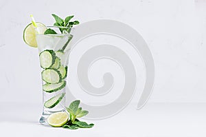 Elegant cocktail with cold detox water with slices cucumber, lime, mint, ice, straw on white wood plank. Refreshing spring healthy