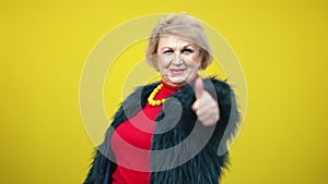 Elegant confident senior woman showing thumb up and winking on yellow background. Portrait of stylish Caucasian lady in