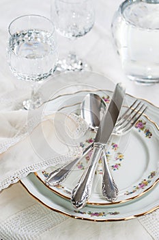 Elegant dinner set with silver knife, fork and spoon lying across luxyry porcelain dishes, and crystal glassware on cream vintage