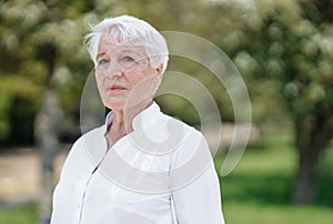 Elegant elderly gray-haired woman in the white shirt is standing in a park on a warm sunny day
