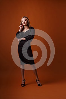 Elegant girl in black dress, on high heel shoes posing on orange background