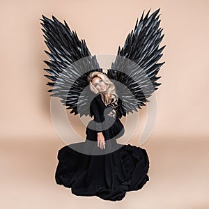 Elegant fashion. Stunning blonde woman in elegant long black dress and big wings on beige background in studio. Luxury evening