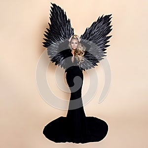 Elegant fashion. Stunning blonde woman in elegant long black dress and big wings on beige background in studio. Luxury evening