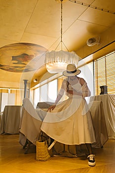 Elegant fashionable woman wearing summer white dress, straw hat, posing in stylish boho interior.