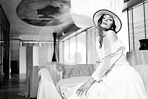 Elegant fashionable woman wearing summer white dress, straw hat, posing in stylish boho interior.