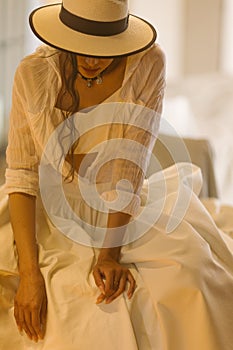 Elegant fashionable woman wearing summer white dress, straw hat, posing in stylish boho interior.