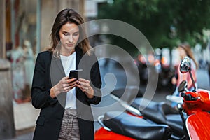 Elegant girl in business suit standing at parking mopeds on street european city and using mobile phone online application rents