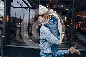 Elegant girl in denim jacket and high heel boots kissing her boyfriend. Handsome man holding his girlfriend while posing