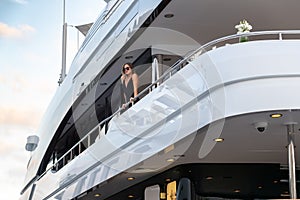 The elegant girl dressed in an evening dress of black color and sunglasses stands on the top deck of a huge yacht in