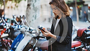 Elegant girl standing at parking mopeds on street city using  smart phone online app rents fast urban transport go work, hipster