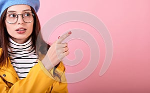 Elegant girl in a stylish beret hat and a yellow cloak in glasses shows a finger to the right on a pink background