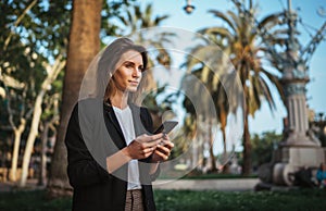 Elegant girl in suit holds a smartphone in hand and looks into distance while walking in a Park in Barcelona, girl ÃÂ¼anager checks