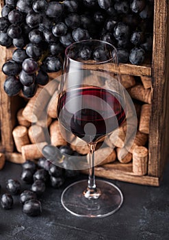 Elegant glass of red wine with dark grapes and corks inside vintage wooden box on black stone background. Natural Light