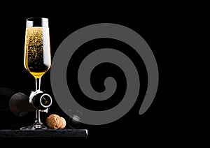 Elegant glass of yellow champagne with cork and wire cage and bottle on black marble board on black background. Space for text