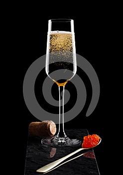 Elegant glass of yellow champagne with red caviar on golden spoon and cork on marble board on black background