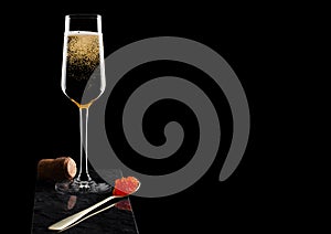Elegant glass of yellow champagne with red caviar on golden spoon and cork on marble board on black background. Space for text