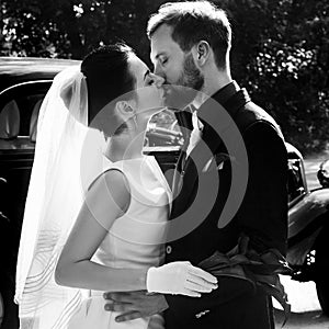 elegant gorgeous bride and handsome stylish groom kissing on background of black car. unusual luxury wedding couple in retro