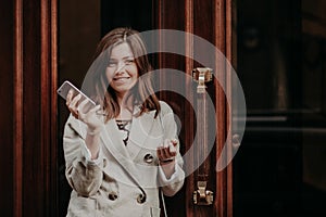 Elegant gorgeous lady in raincoat, holds mobile phone, waits for call, poses outdoor near doors, going for work, feels pleased, up