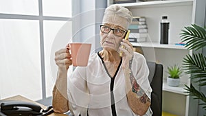 Elegant grey-haired senior woman boss engrossed in a serious phone conversation, relaxedly sipping coffee at her office