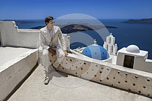 Elegant handsome man in suit is posing near Church of the Three Bells in Fira in Santorini