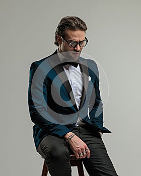 elegant high class man with glasses looking down and sitting with hand in pocket