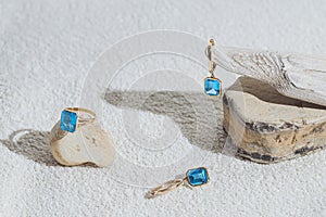 Elegant jewellery set of gold earrings and ring with blue topaz on white background with stones and wood. Minimal style