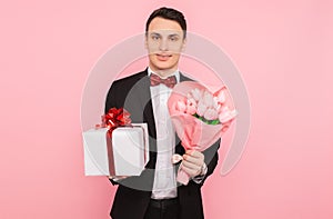 Elegant man, in a suit, with a bouquet of flowers, and a gift box, on a pink background, the concept of women`s day