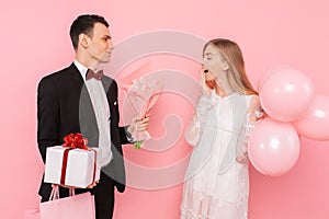 Elegant man in a suit, gives a box with a gift and a bouquet of flowers, to a beautiful woman, on a pink background. Women`s day