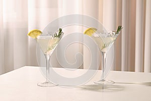 Elegant martini glasses with fresh cocktail, rosemary and lemon slices on white table indoors