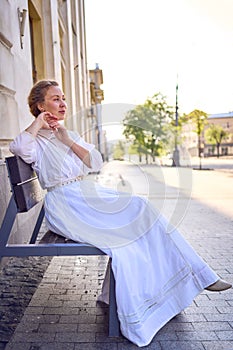 elegant middle age woman in a white vintage dress sitting on a bench in the morning city