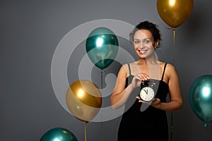 Elegant mixed race woman dressed in evening dress, holds an alarm clock and cute smiles with toothy smile looking at camera,