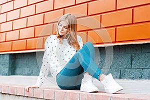 Elegant modern young cheerful woman a blonde in a white sweater in trendy jeans in sneakers sits on the ground near