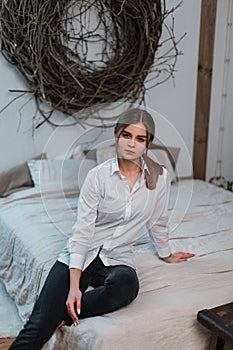 Elegant modern young woman in a white fashionable elegant shirt in stylish black jeans sits on a bed in a vintage room. Pretty