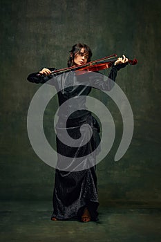 Elegant musician, beautiful young woman in black dress playing violin against dark green vintage background. Symphony